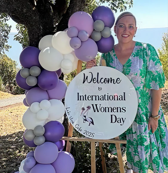 Woman Standing Beside An International Women's Day Welcome Sign With Purple Balloon Display